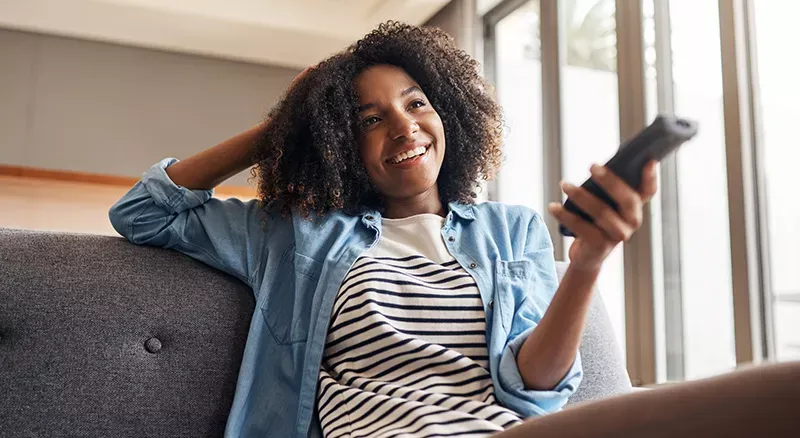 Woman sits on grey couch while pointing remote towards TV